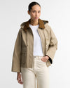 A woman modeling a beige hooded jacket with a brown suede-like collar, worn over a white t-shirt and cream-colored pants, against a plain white studio background.