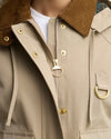 Close-up of a beige hooded jacket with a brown corduroy collar, gold-tone snap buttons, a central zipper with a ring pull, and a matching gold D-ring detail on the chest.