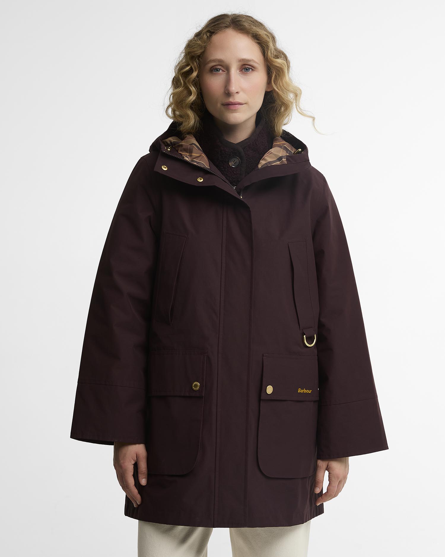 A woman wearing a dark burgundy hooded jacket with a plaid-lined hood, gold-tone hardware, and front flap pockets, photographed against a plain light gray studio background.