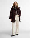 A woman stands against a light gray studio background wearing a dark burgundy hooded raincoat over a black-and-white striped long-sleeve top, cream wide-leg trousers, and black shoes.