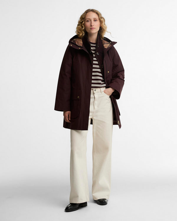 A woman stands against a light gray studio background wearing a dark burgundy hooded raincoat over a black-and-white striped long-sleeve top, cream wide-leg trousers, and black shoes.