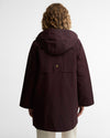 A person viewed from the back wearing a hooded jacket in a deep burgundy or maroon shade, paired with light beige pants, against a plain light gray studio background. A single gold-colored button detail is centered on the back.