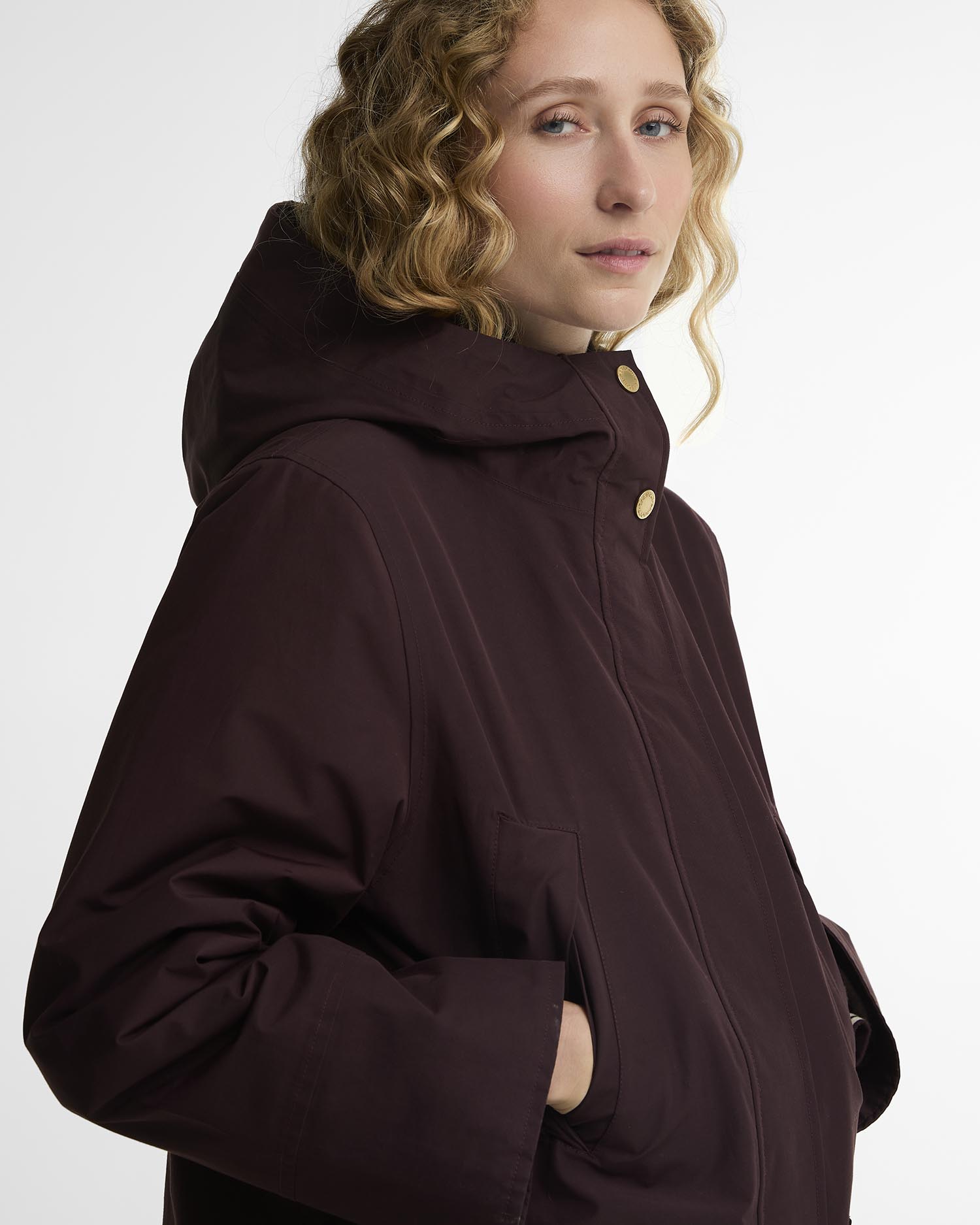 A woman wearing a dark plum-brown hooded jacket with gold-tone snap buttons, shown in a close-up side profile against a clean white studio background.