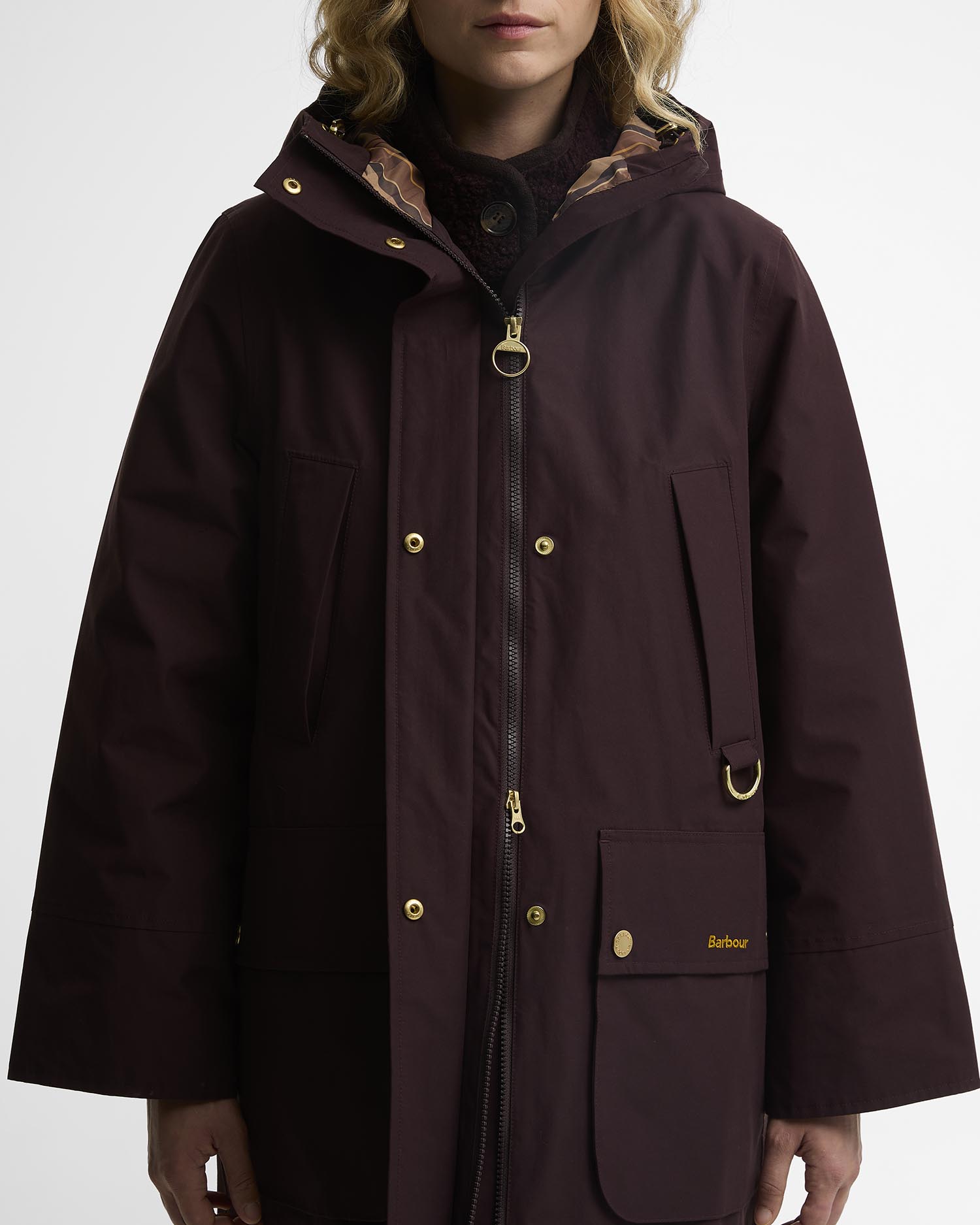 A model wearing a Barbour hooded raincoat in deep burgundy. The jacket features gold-tone metal hardware, including snap buttons, a two-way zipper pull, and a Barbour logo on the front pocket, set against a plain light gray background.