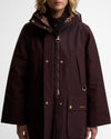 A model wearing a Barbour hooded raincoat in deep burgundy. The jacket features gold-tone metal hardware, including snap buttons, a two-way zipper pull, and a Barbour logo on the front pocket, set against a plain light gray background.