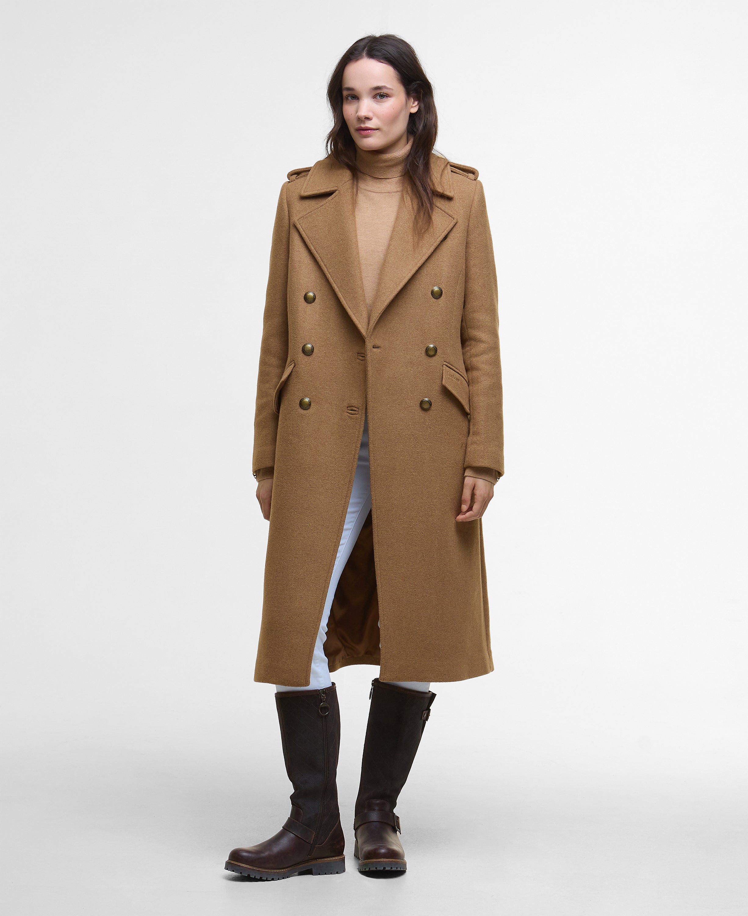 A woman modeling a long camel-brown double-breasted coat over a beige turtleneck, light blue jeans, and dark brown knee-high boots, photographed against a plain light gray studio background.