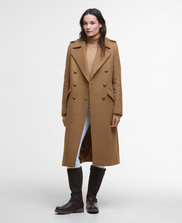A woman modeling a long camel-brown double-breasted coat over a beige turtleneck, light blue jeans, and dark brown knee-high boots, photographed against a plain light gray studio background.