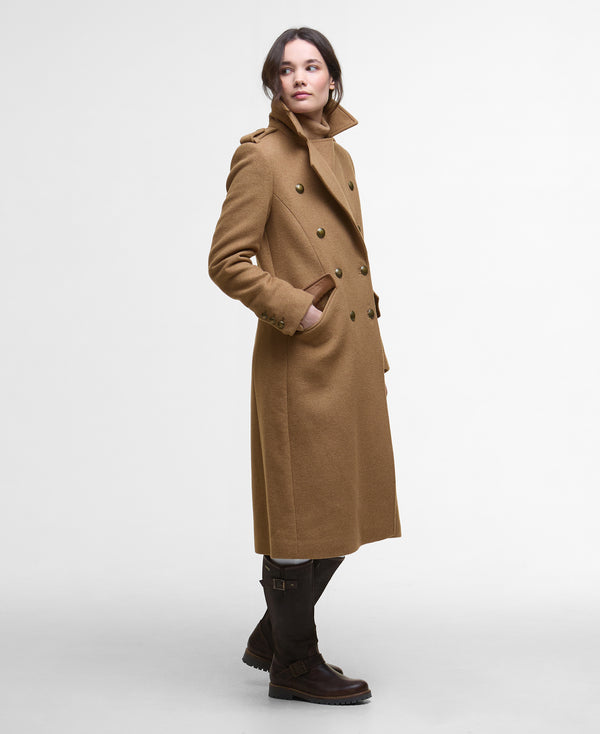A woman modeling a mid-length camel brown double-breasted wool coat with a high collar, paired with dark brown leather boots, against a plain light gray studio background.