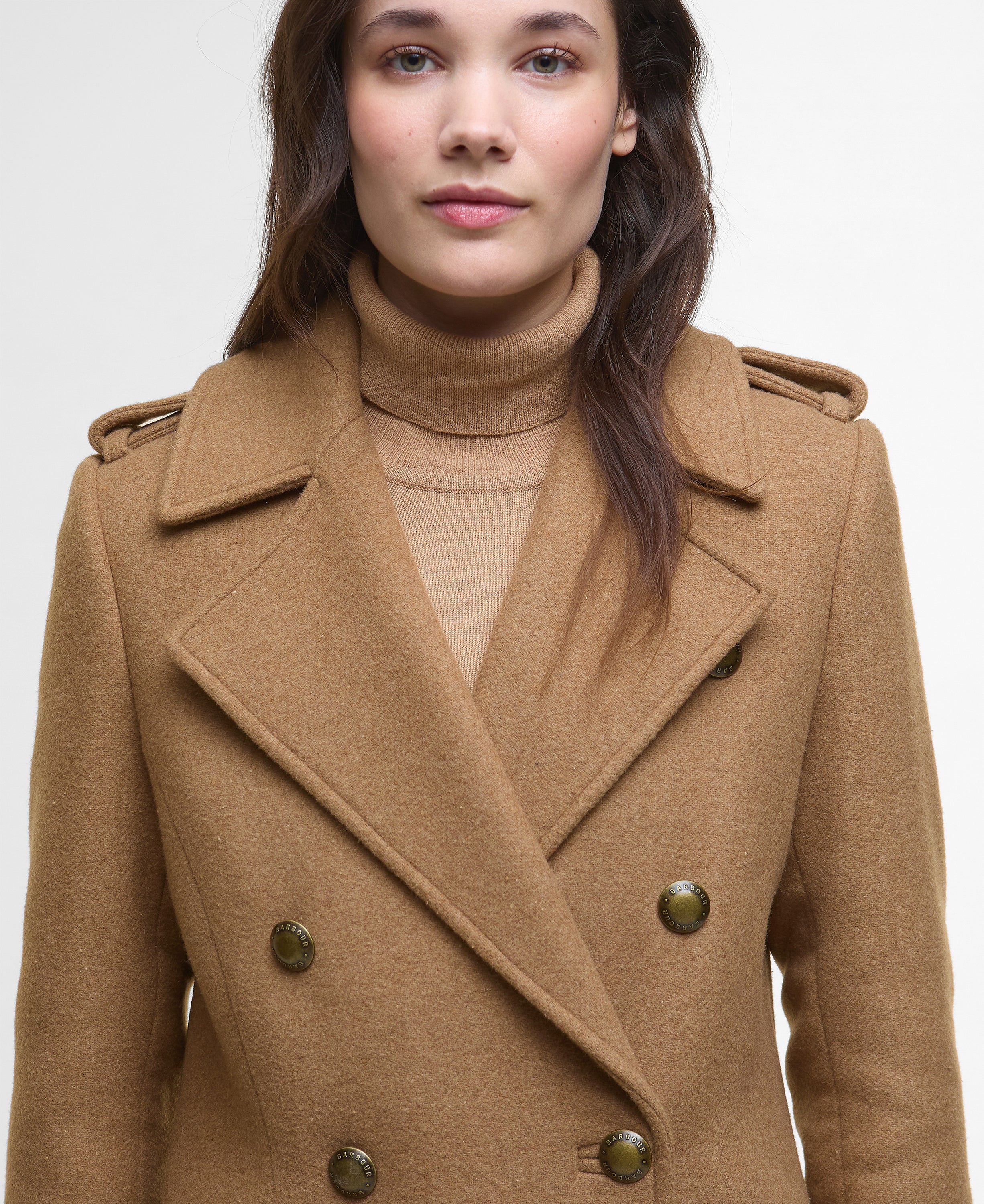 A woman wearing a camel brown double-breasted coat over a matching beige turtleneck sweater, photographed against a clean white studio background.