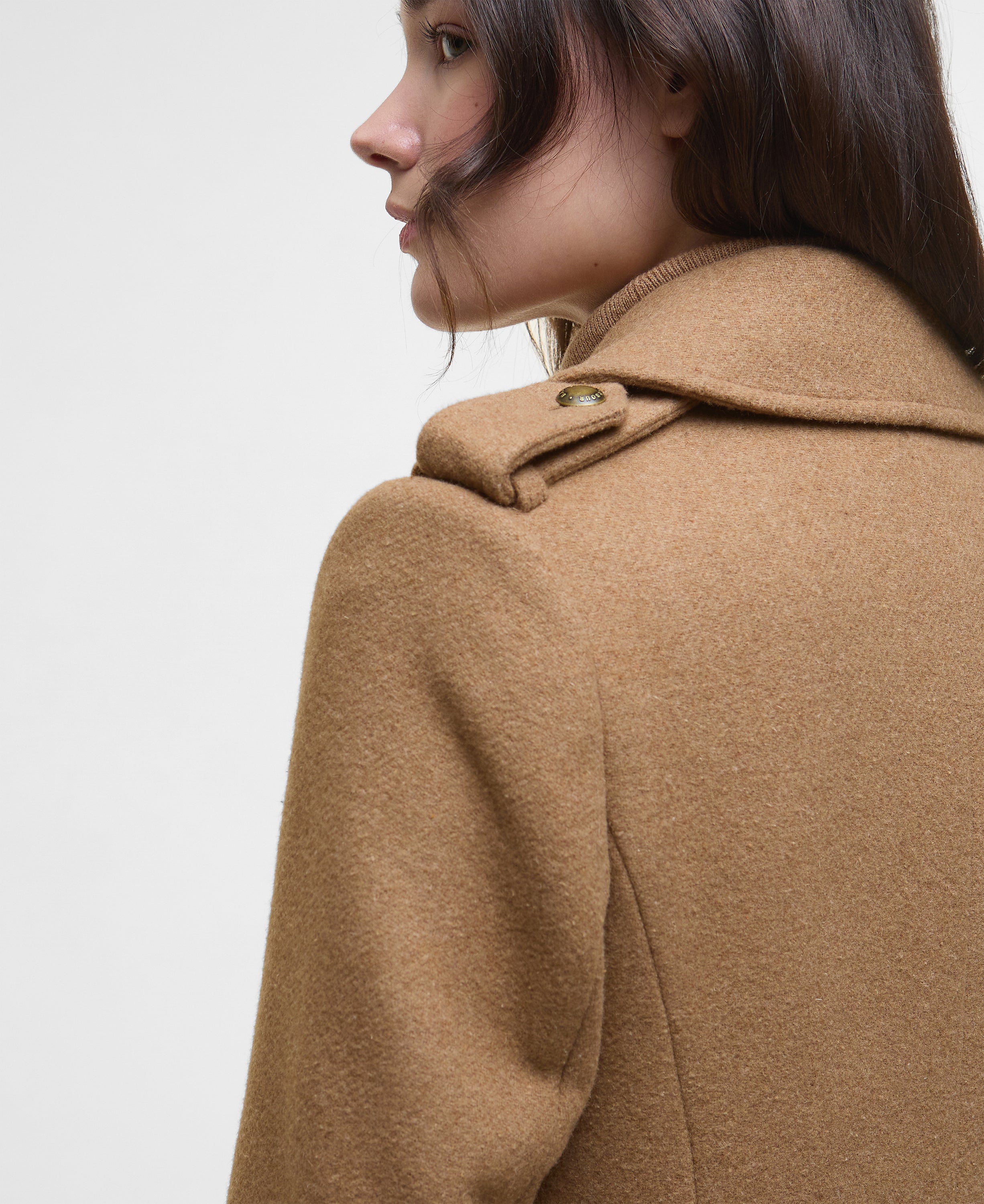 Close-up side profile of a model wearing a camel brown wool coat with a structured collar and a single decorative button, set against a plain light gray background.
