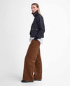 A woman stands in profile wearing a black jacket with gold-tone hardware, a white shirt tucked into wide-leg brown corduroy pants, and black shoes, against a clean white studio background.