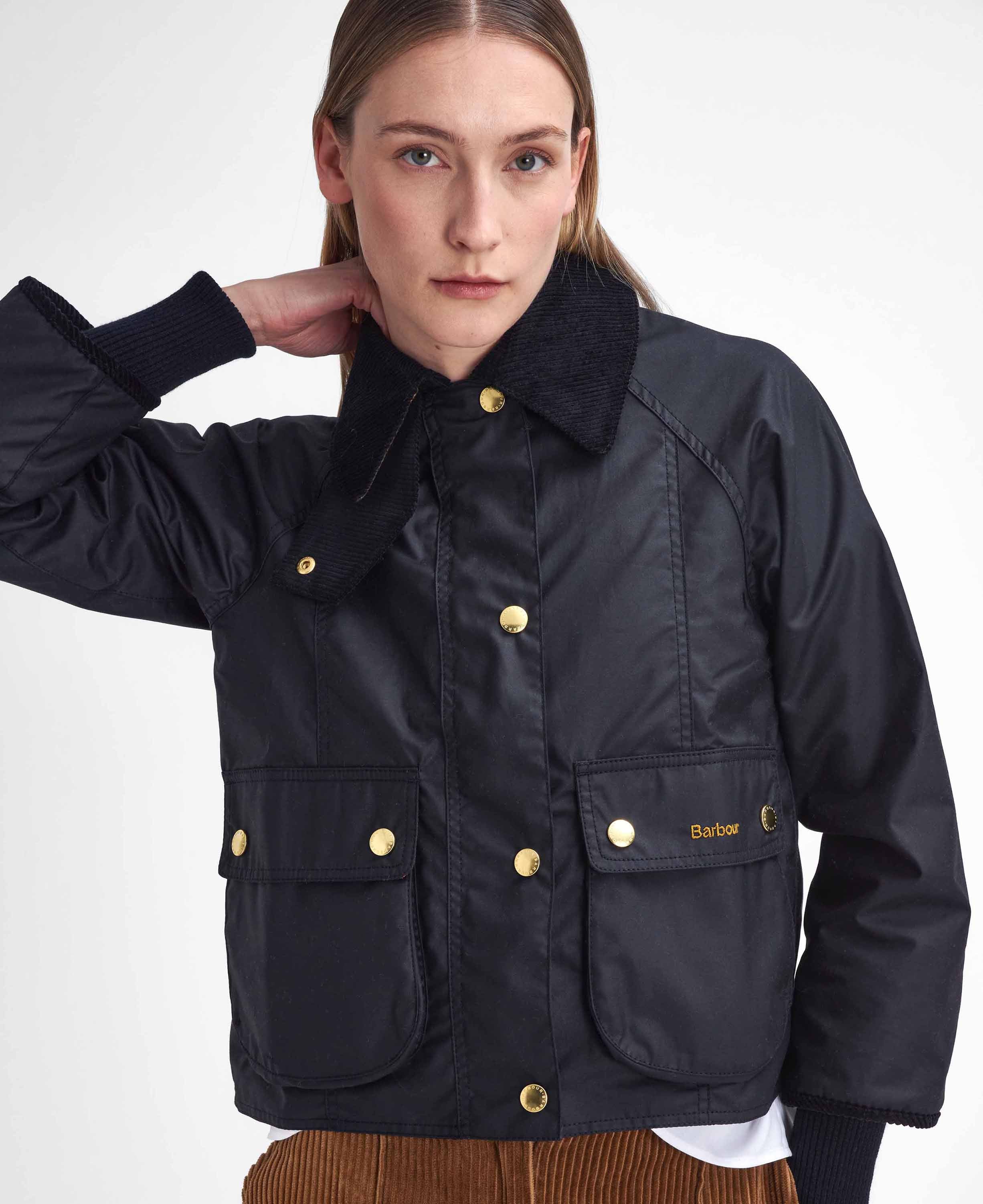A woman wearing a black Barbour waxed cotton jacket with gold buttons and a dark corduroy collar posed against a light gray studio background.