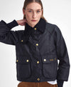 A woman wearing a black Barbour waxed cotton jacket with gold buttons and a dark corduroy collar posed against a light gray studio background.