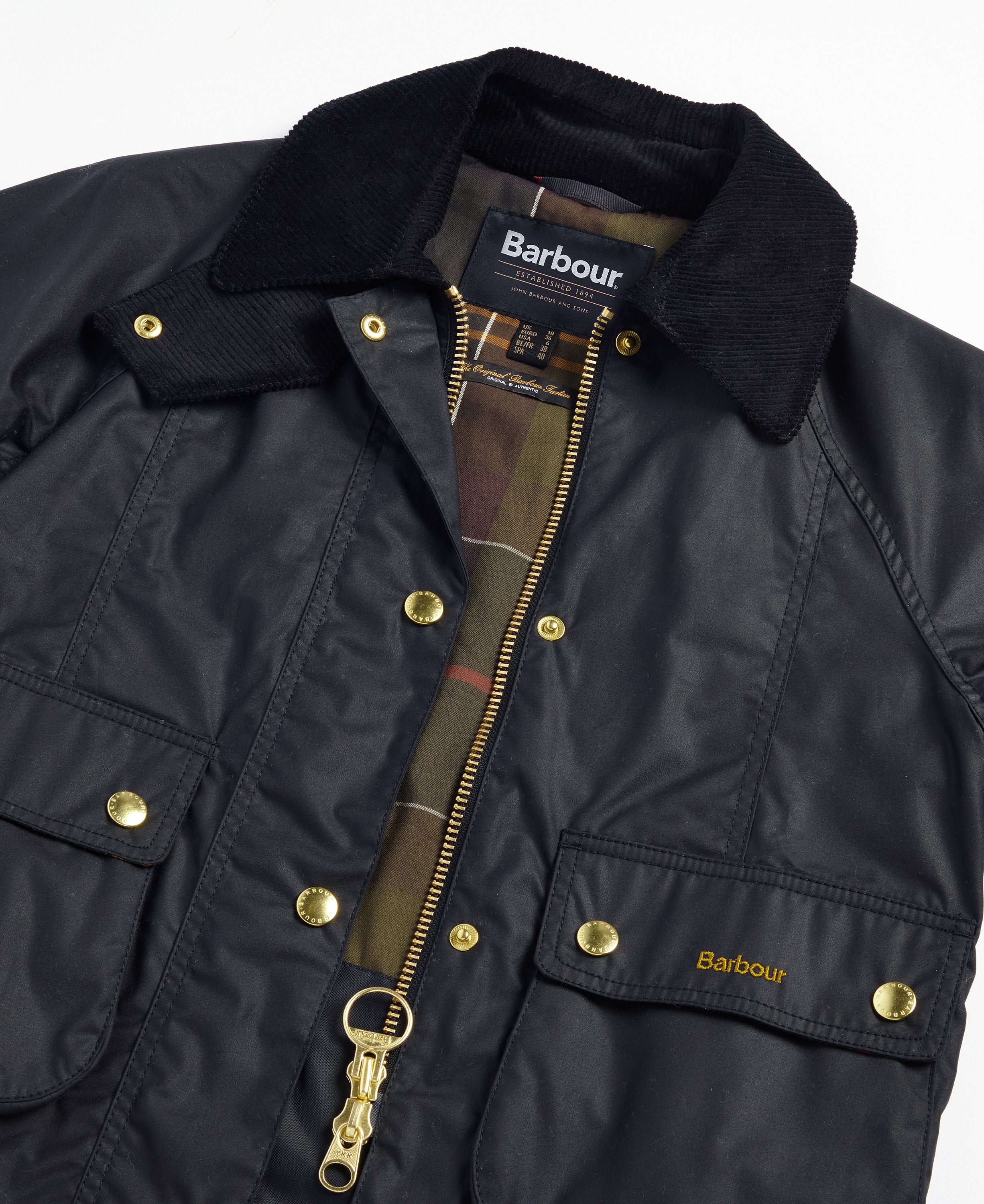 Close-up of a Barbour waxed jacket in black, featuring a black corduroy collar, gold-tone snap and zipper hardware, and a plaid lining visible at the open collar.