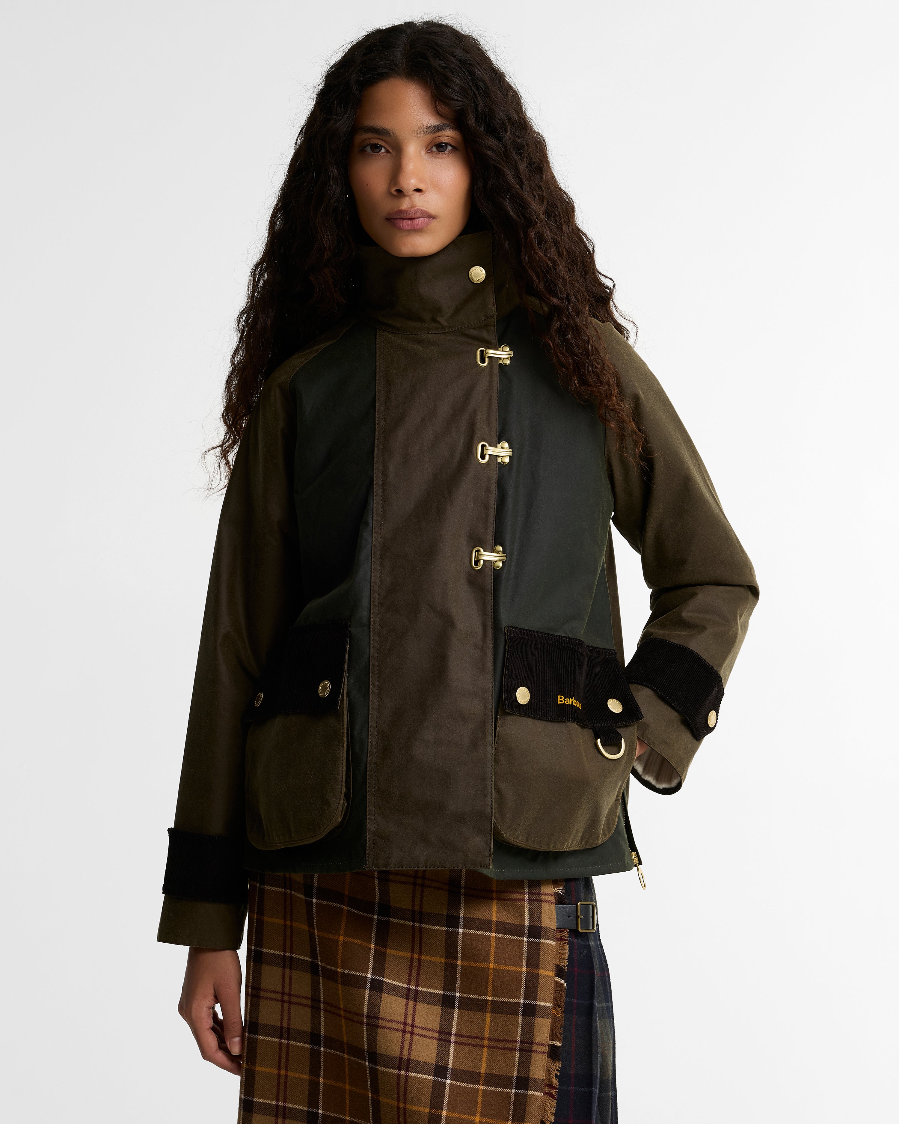 Model wearing the Barbour Hazelwood Wax Jacket, showing the two-tone waxed cotton paneling, gold equestrian hook-and-eye closures, and corduroy patch pockets with Barbour script, styled over a brown tartan midi skirt.