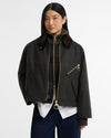 A model wearing a dark gray plaid waxed cotton jacket with a brown collar, gold-tone zippers, and a white shirt tucked into dark blue jeans, shot against a light gray studio background.