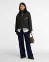 A model stands against a light gray studio background wearing a dark cropped plaid waxed cotton jacket with a corduroy collar, navy wide-leg trousers, a white shirt, and holding a brown plaid handbag.