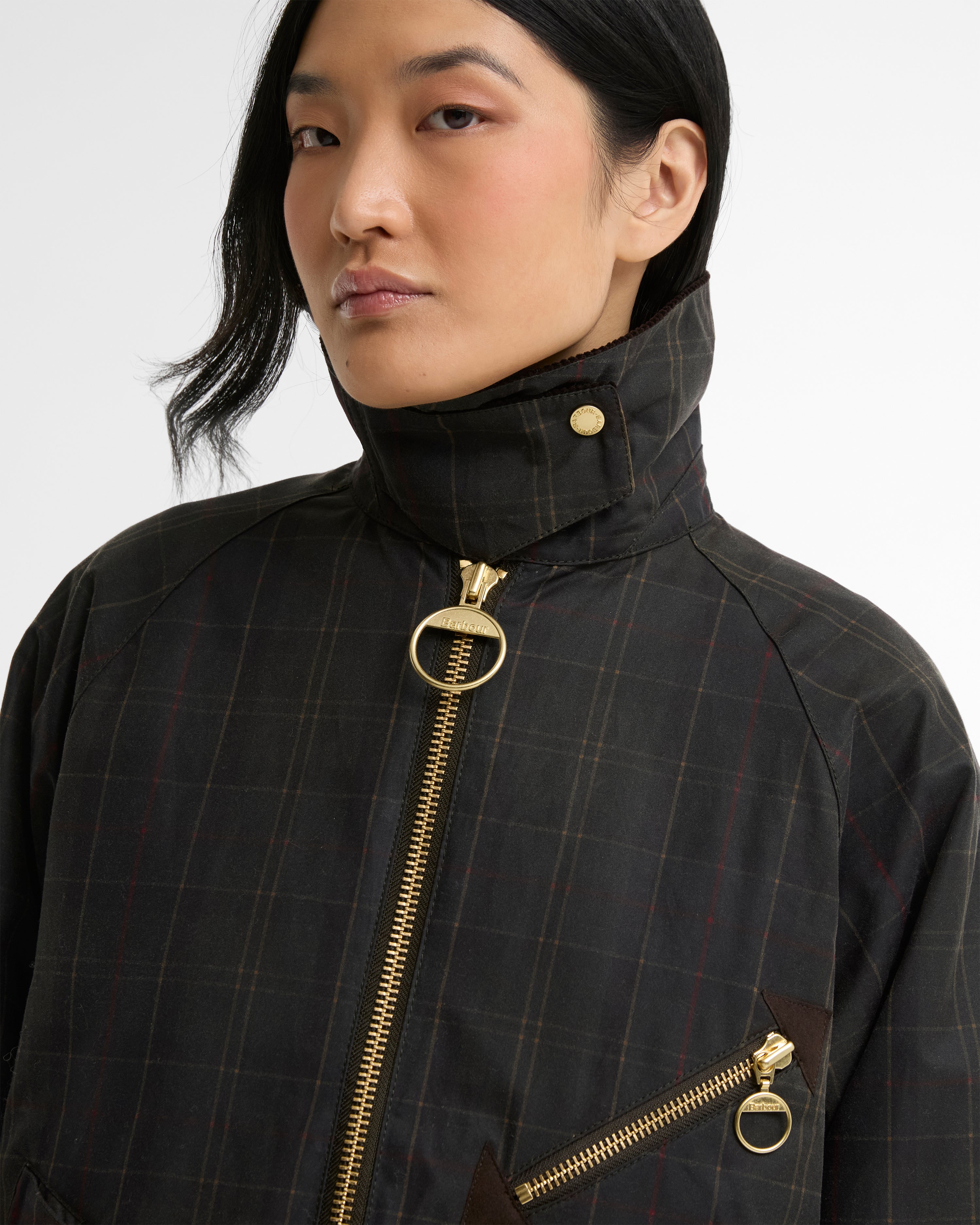 A close-up of a model wearing a dark charcoal gray plaid waxed cotton jacket with prominent gold-tone hardware, including a large ring pull on the front zipper and a smaller ring pull on the chest pocket.