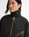 A close-up of a model wearing a dark charcoal gray plaid waxed cotton jacket with prominent gold-tone hardware, including a large ring pull on the front zipper and a smaller ring pull on the chest pocket.