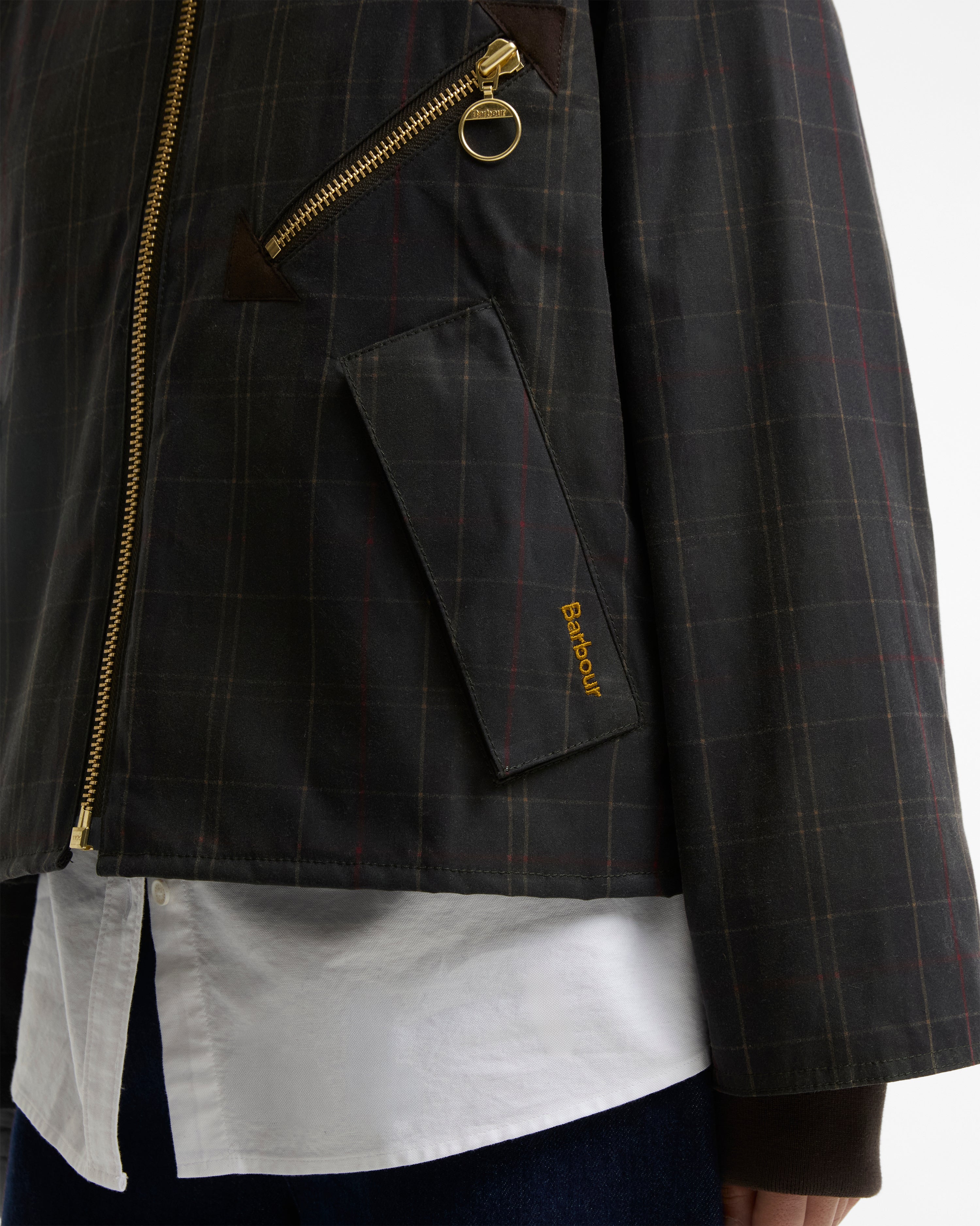 Close-up of a dark gray plaid jacket with gold zippers, featuring a diagonal zip pocket with a gold ring pull and a small gold Barbour logo on the pocket flap, worn over a white shirt.