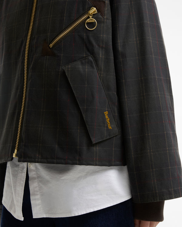 Close-up of a dark gray plaid jacket with gold zippers, featuring a diagonal zip pocket with a gold ring pull and a small gold Barbour logo on the pocket flap, worn over a white shirt.