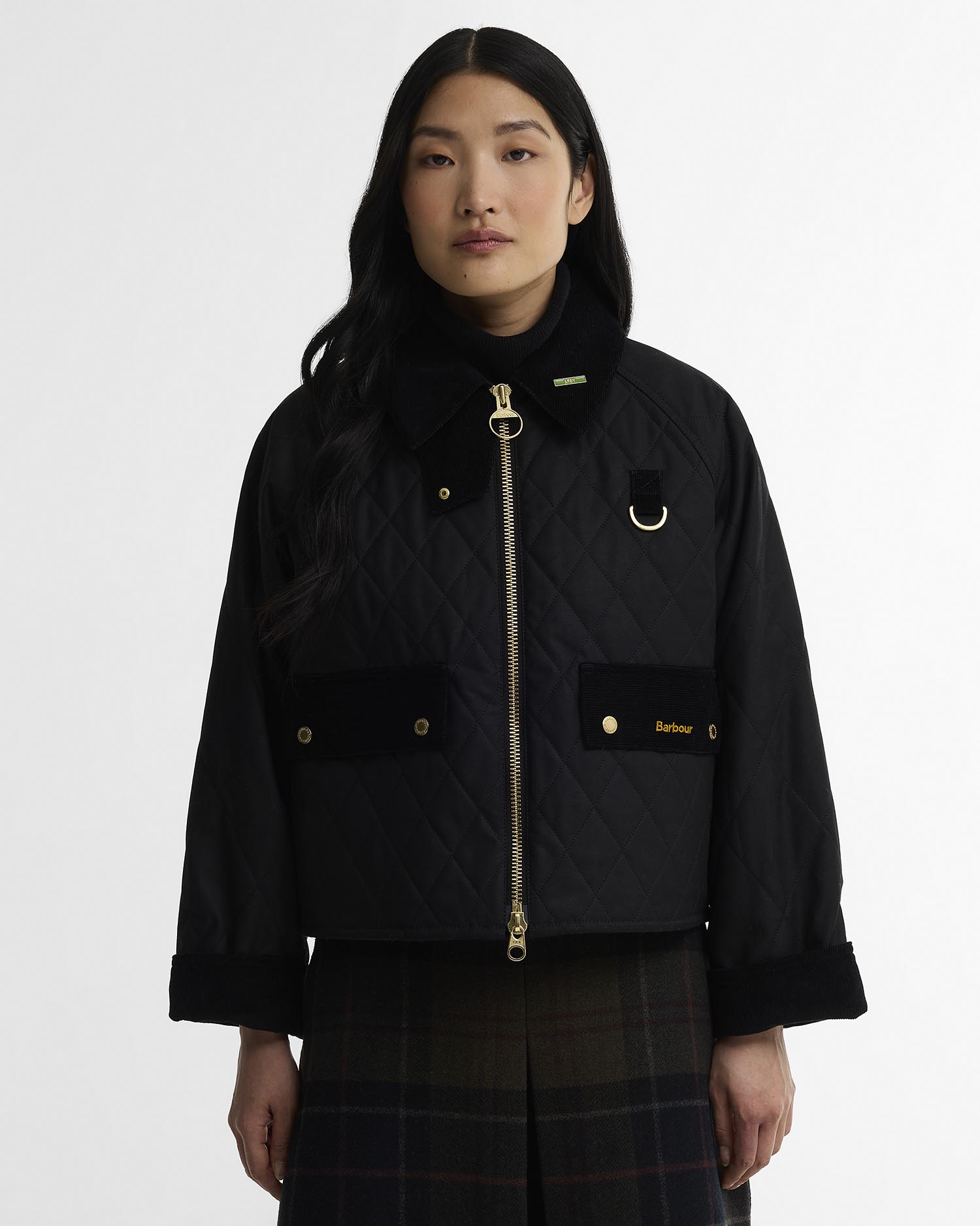 A model wearing a black Barbour quilted jacket with gold-tone hardware and a black turtleneck underneath, set against a plain light gray studio background.
