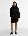 A model stands against a light gray studio background wearing a black jacket with gold-toned hardware and a dark plaid mini skirt.