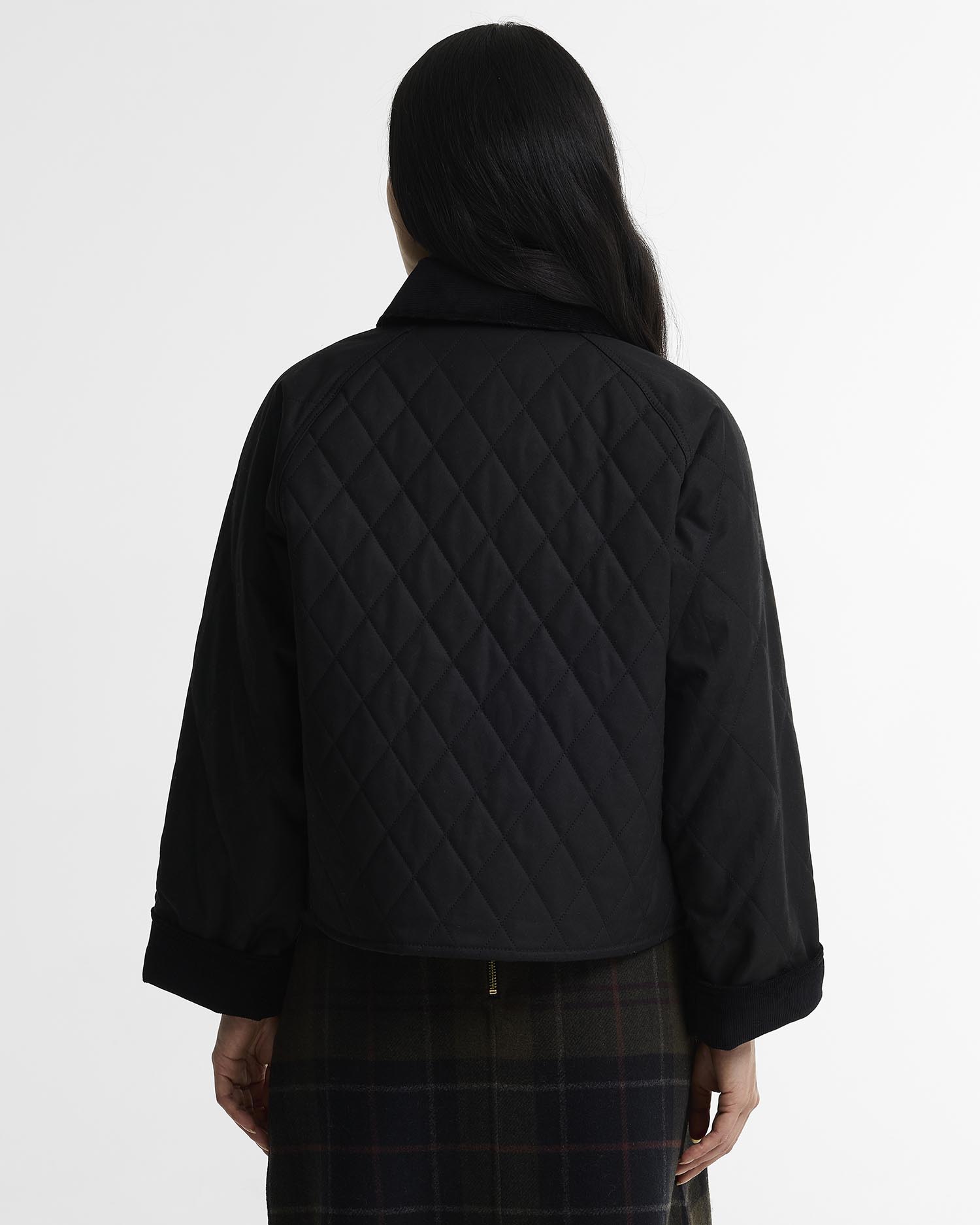 A model shown from the back wearing a black quilted cropped jacket with a high collar and a dark plaid skirt, against a plain white studio background.