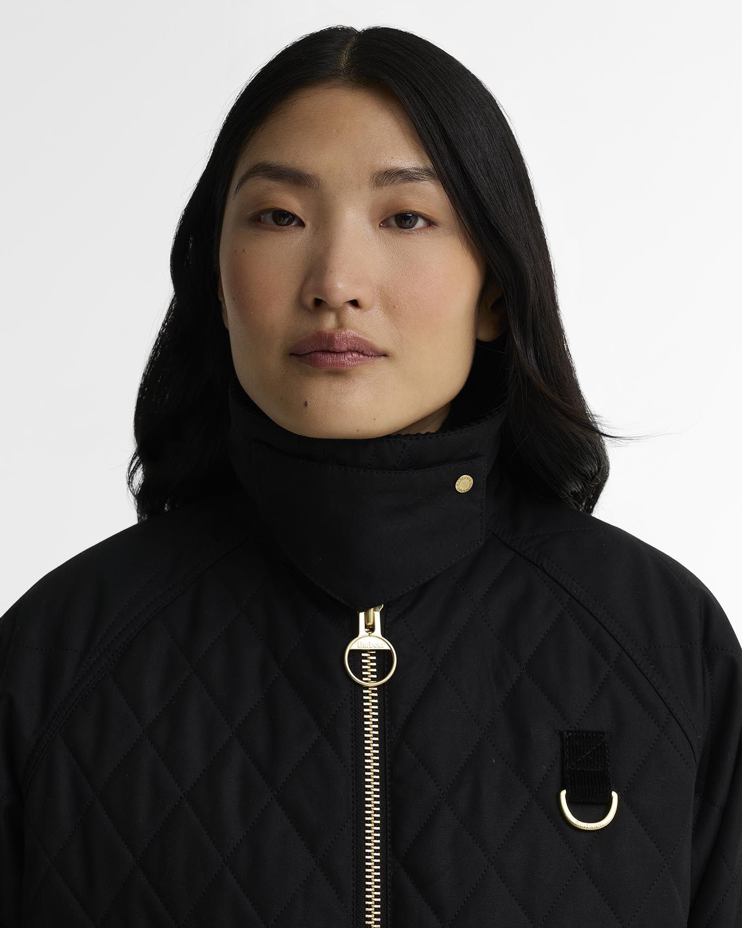 A close-up portrait of a model wearing a black quilted jacket with gold-tone hardware, including a zipper, ring pull, and chest D-ring, against a plain light gray background.