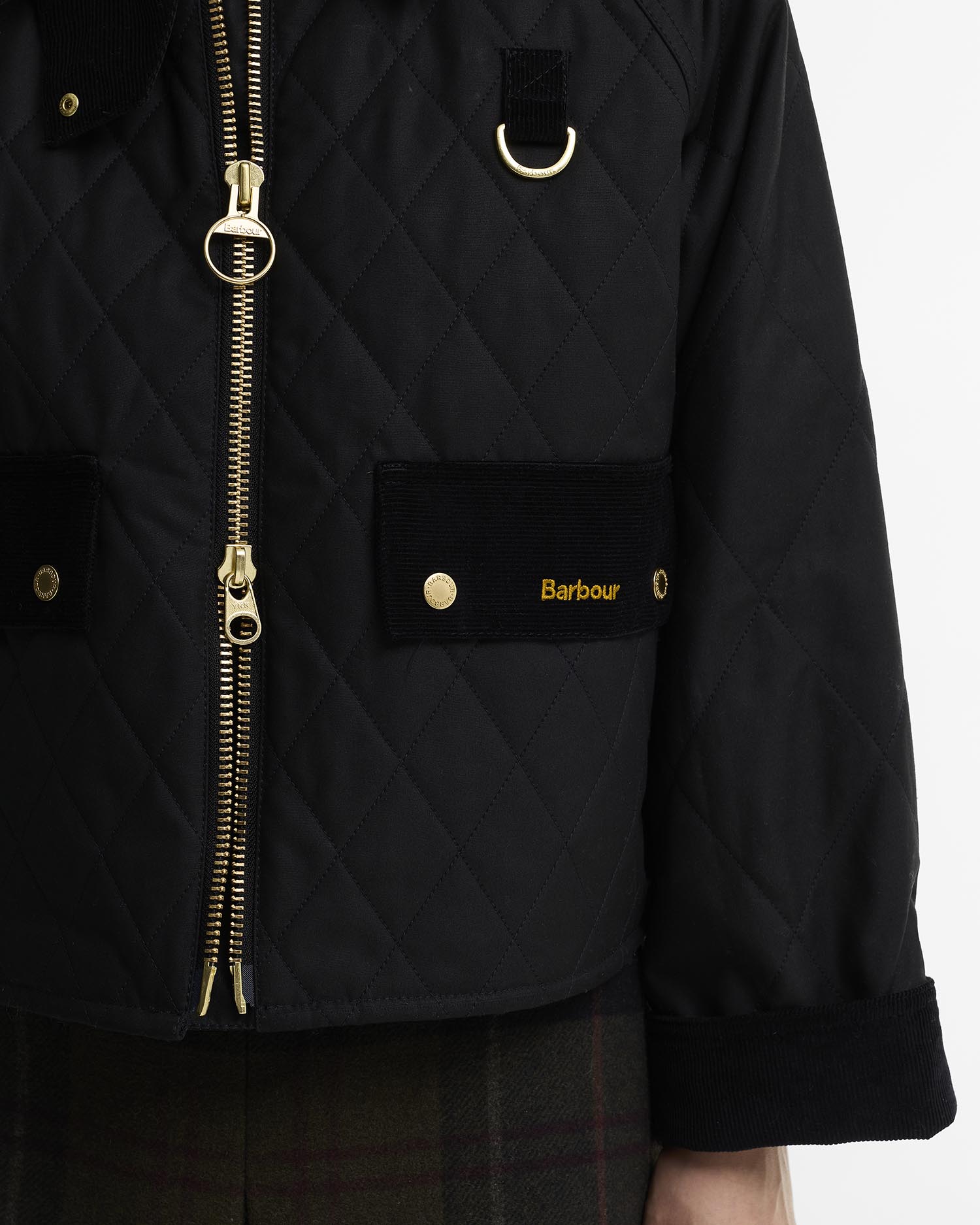 Close-up of a black quilted wax jacket with gold-tone hardware, including a two-way front zipper, D-ring detail, and a patch pocket with the Barbour logo.
