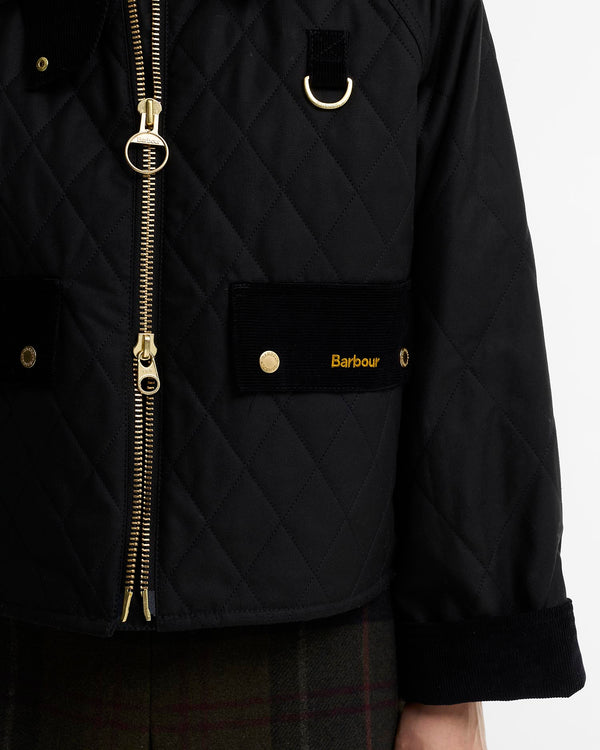 Close-up of a black quilted wax jacket with gold-tone hardware, including a two-way front zipper, D-ring detail, and a patch pocket with the Barbour logo.