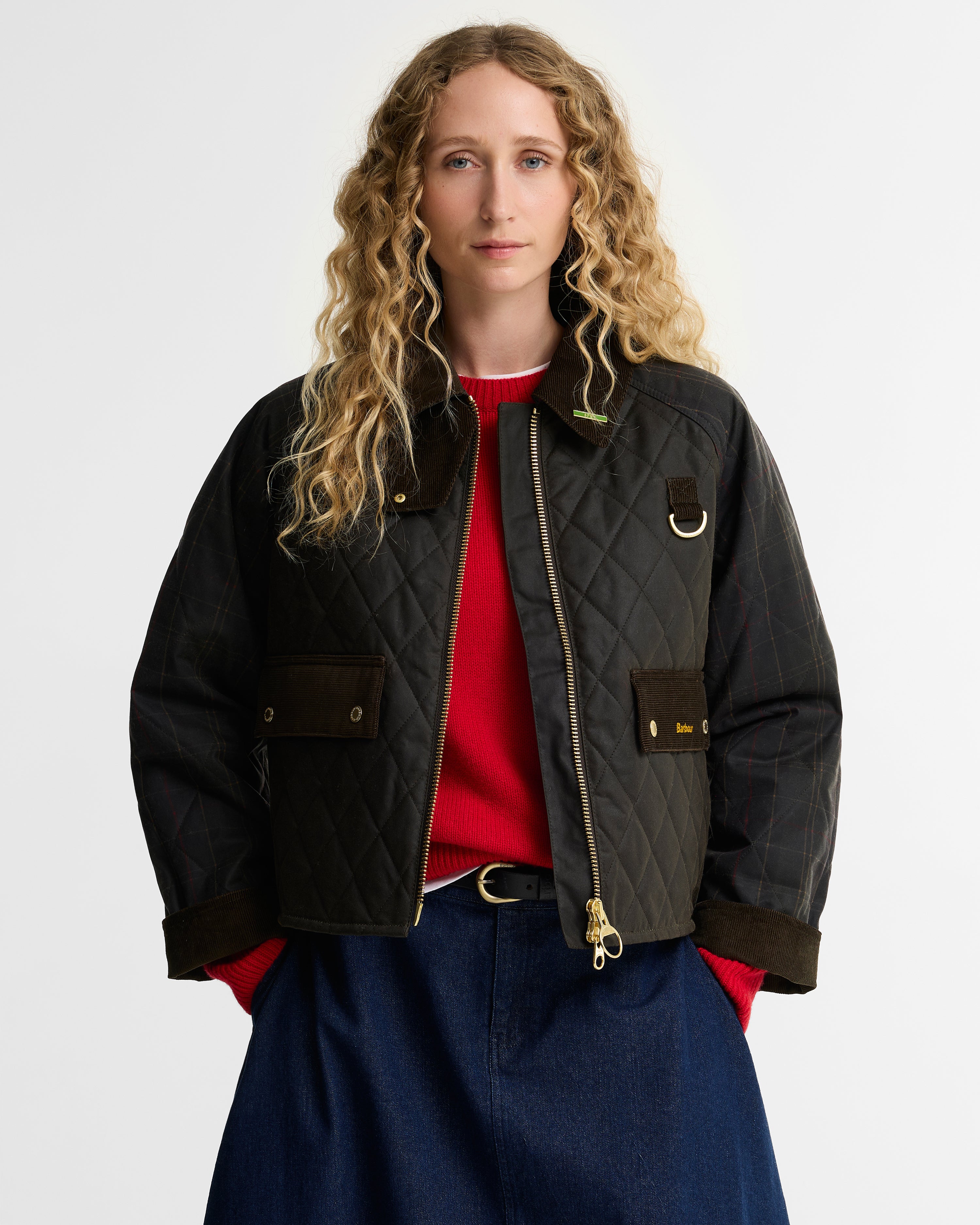 A woman with long curly blonde hair models a dark olive quilted bomber jacket with brown trim, worn over a red top and blue jeans against a light gray studio background.
