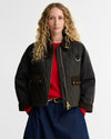 A woman with long curly blonde hair models a dark olive quilted bomber jacket with brown trim, worn over a red top and blue jeans against a light gray studio background.