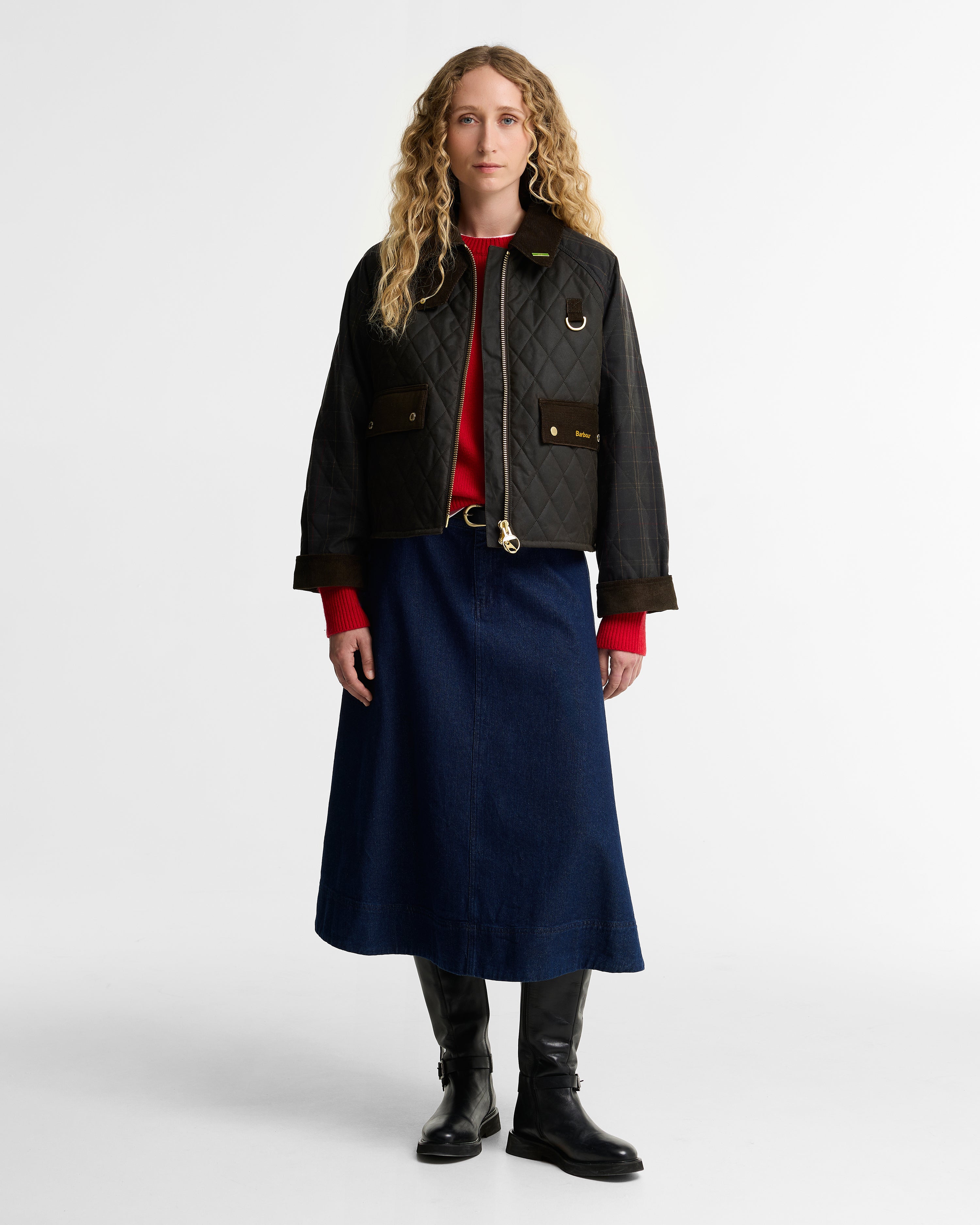 A model stands against a light gray studio background wearing a dark olive quilted waxed jacket with brown trim over a red top, a navy midi skirt, and black boots.