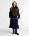 A model stands against a light gray studio background wearing a dark olive quilted waxed jacket with brown trim over a red top, a navy midi skirt, and black boots.