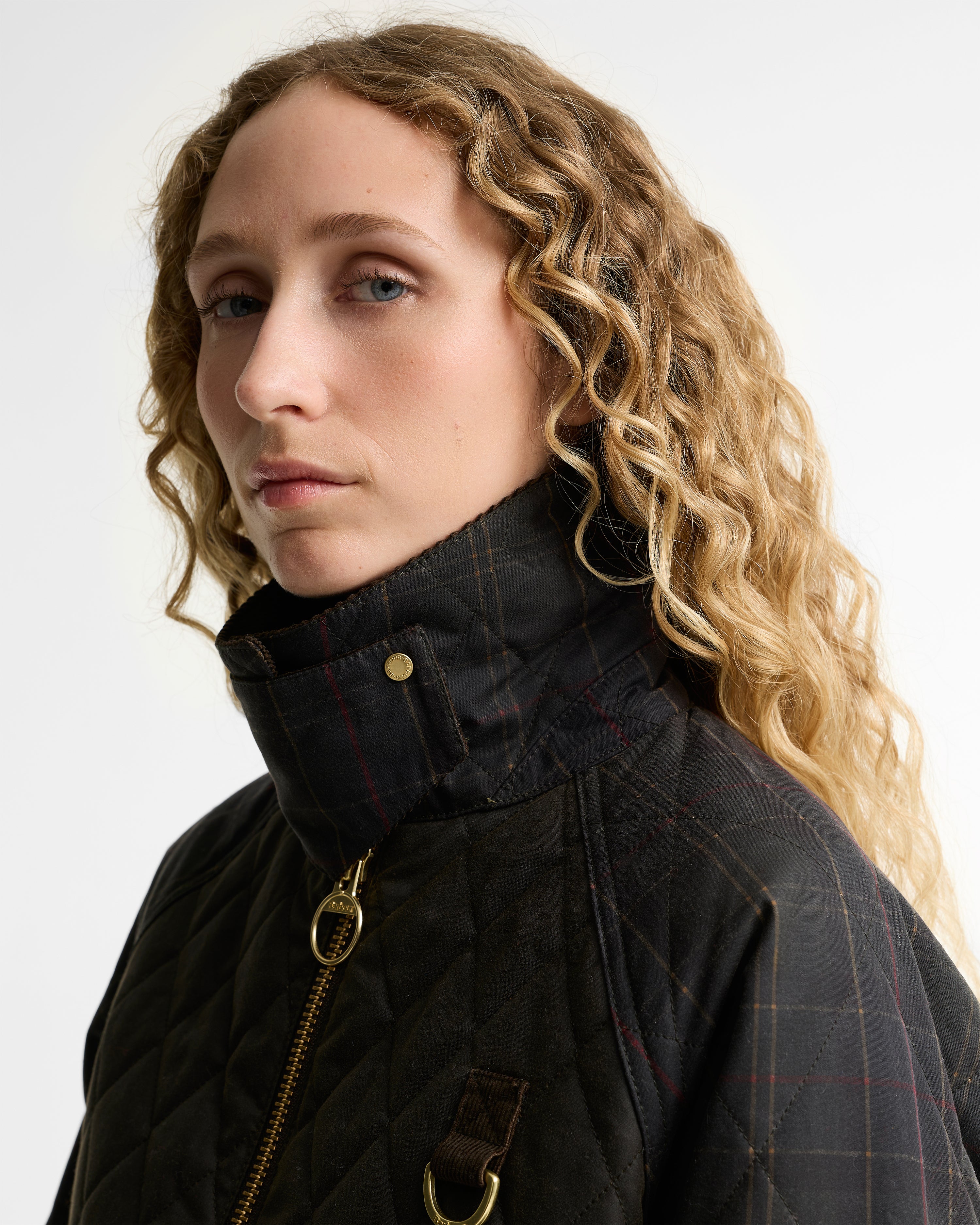 A close-up portrait of a woman with curly blonde hair wearing a dark olive quilted waxed jacket with a high collar, gold zipper, and subtle red windowpane plaid pattern, set against a plain light gray background.