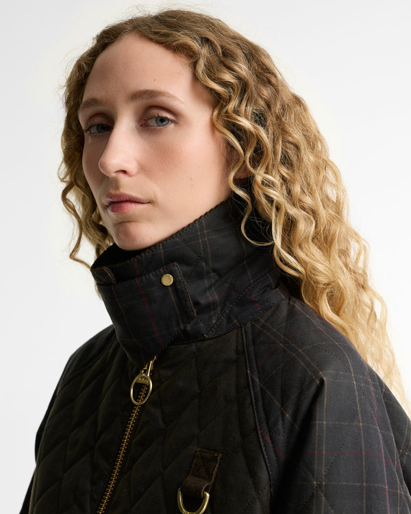 A close-up portrait of a woman with curly blonde hair wearing a dark olive quilted waxed jacket with a high collar, gold zipper, and subtle red windowpane plaid pattern, set against a plain light gray background.