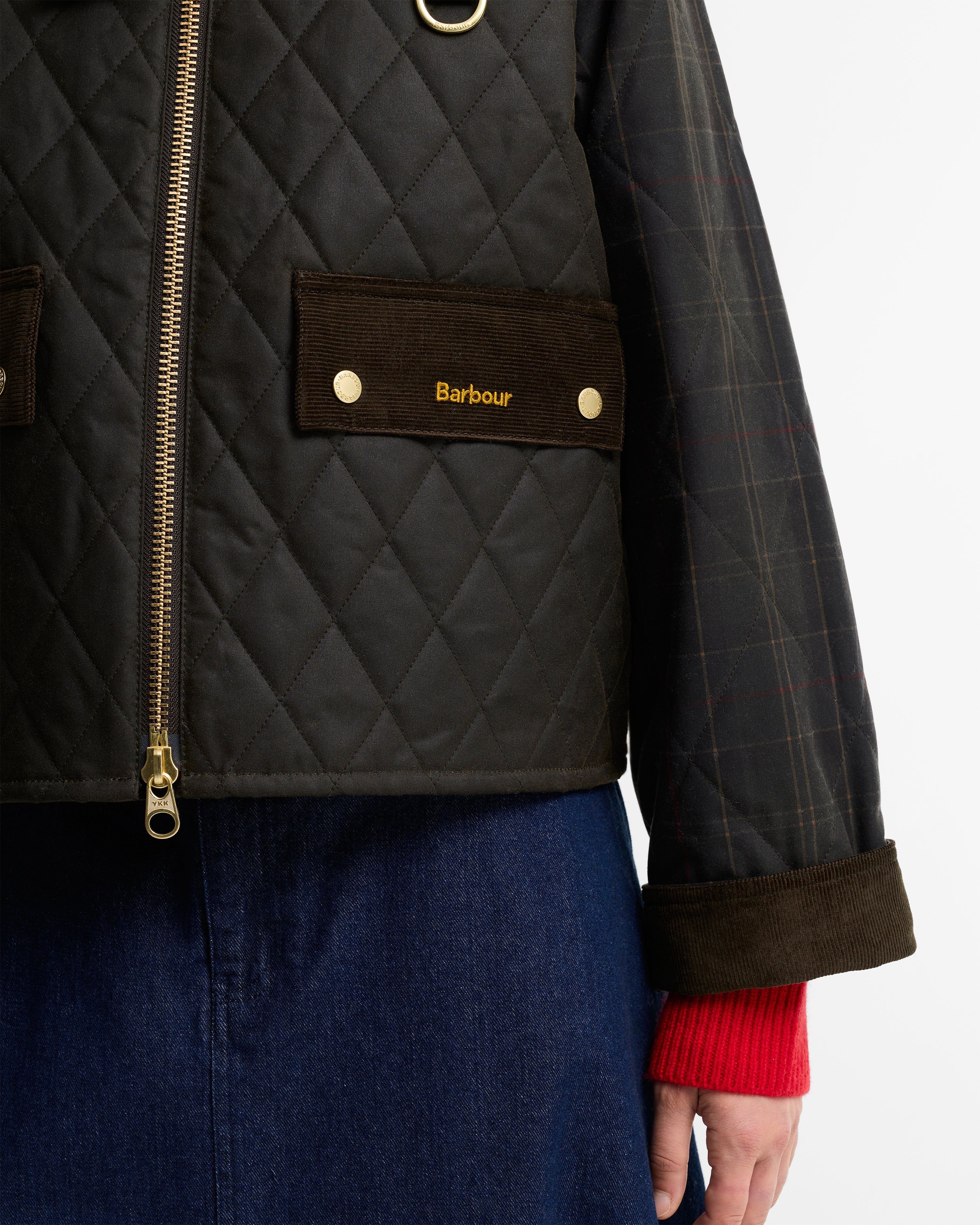 Close-up of a Barbour quilted jacket with a diamond-quilted dark olive body, brown corduroy pocket flap with gold logo, gold zipper, and a plaid sleeve, worn over a red knit cuff and blue skirt.