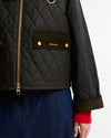Close-up of a Barbour quilted jacket with a diamond-quilted dark olive body, brown corduroy pocket flap with gold logo, gold zipper, and a plaid sleeve, worn over a red knit cuff and blue skirt.