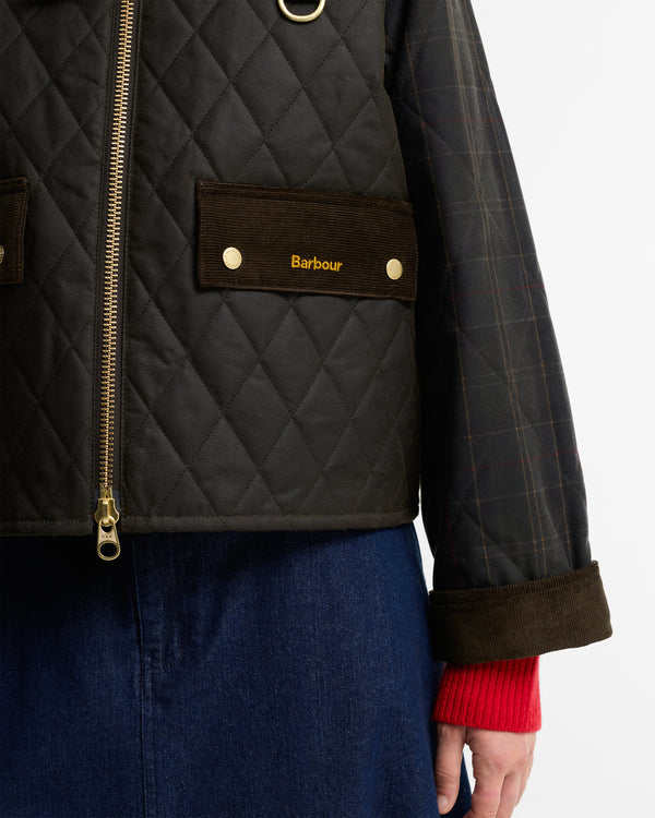 Close-up of a Barbour quilted jacket with a diamond-quilted dark olive body, brown corduroy pocket flap with gold logo, gold zipper, and a plaid sleeve, worn over a red knit cuff and blue skirt.