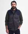 A man wearing a dark navy quilted puffer vest over a dark green knit sweater and dark jeans, standing against a plain light gray studio background.