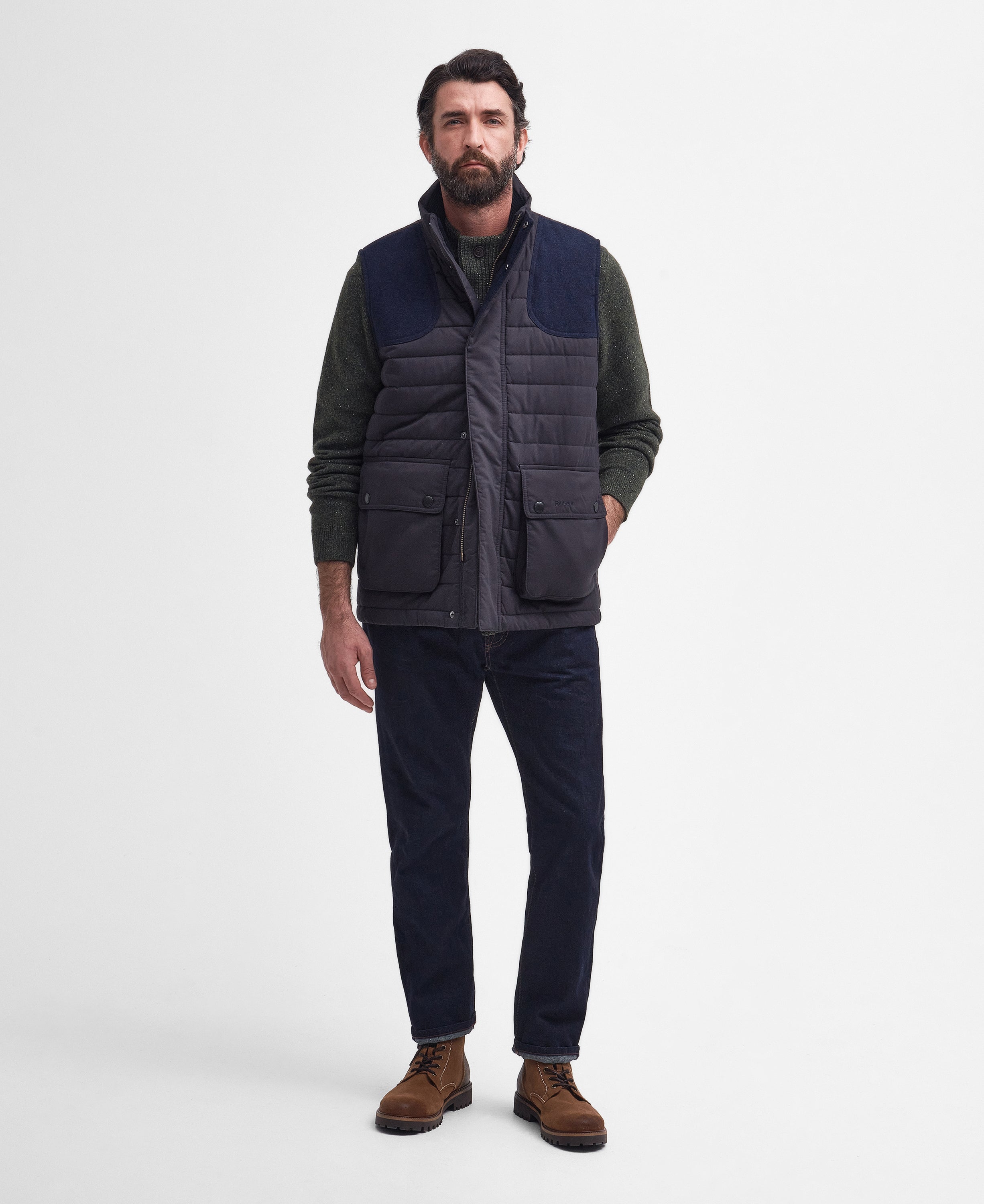 A man wearing a dark navy quilted puffer vest over a dark green long-sleeve shirt, paired with dark navy trousers and brown suede boots, standing against a light gray studio background.