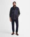 A man wearing a dark navy quilted puffer vest over a dark green long-sleeve shirt, paired with dark navy trousers and brown suede boots, standing against a light gray studio background.