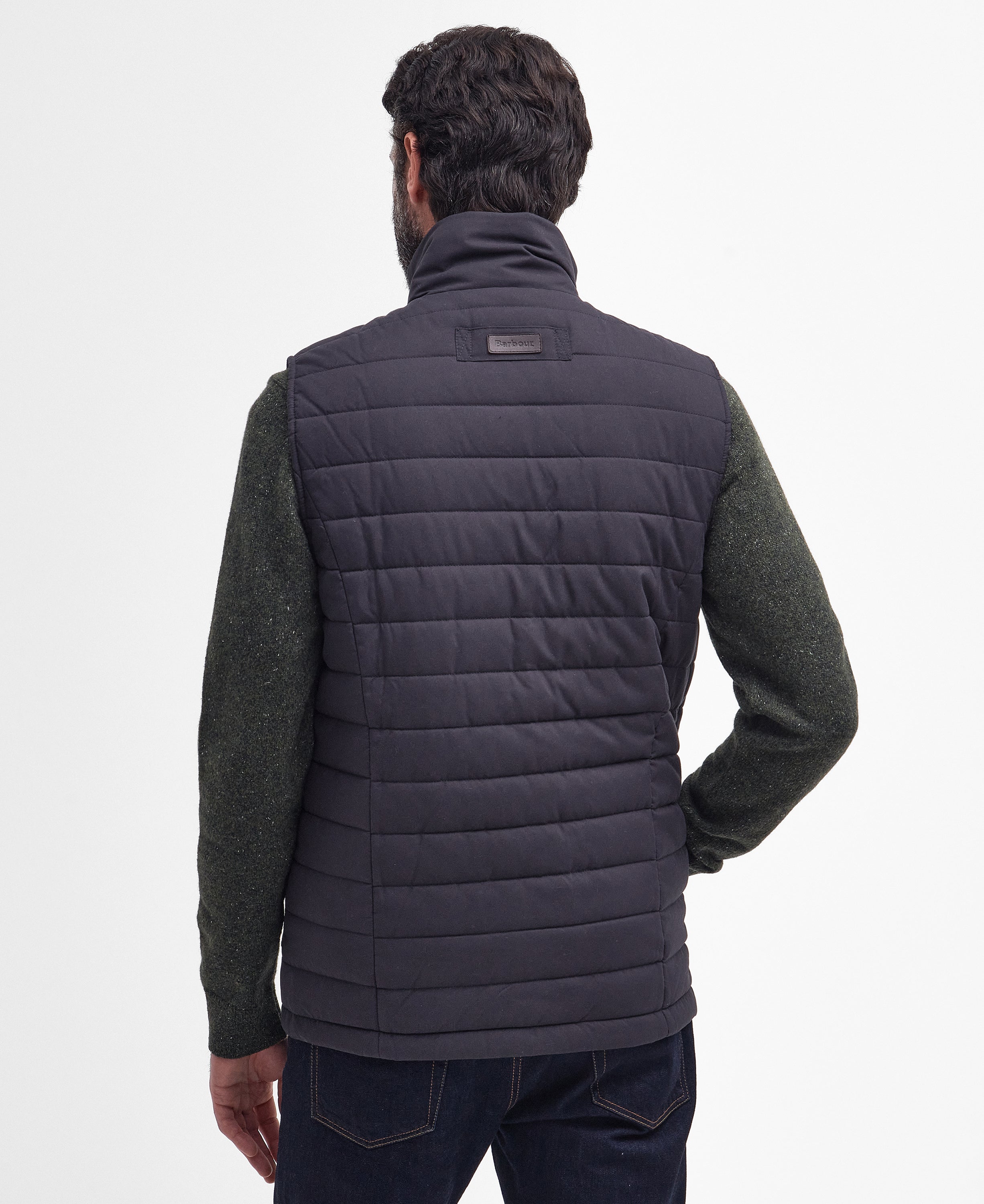 A man viewed from the back wearing a dark navy quilted puffer vest over a dark green knit sweater and dark blue jeans, against a plain white studio background.