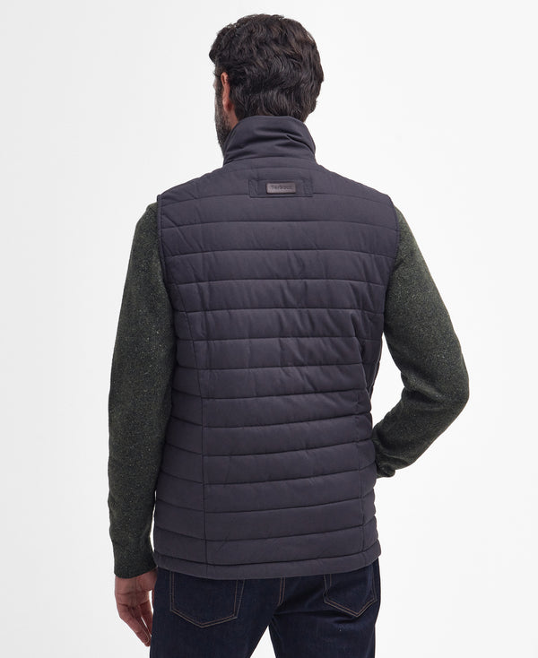 A man viewed from the back wearing a dark navy quilted puffer vest over a dark green knit sweater and dark blue jeans, against a plain white studio background.