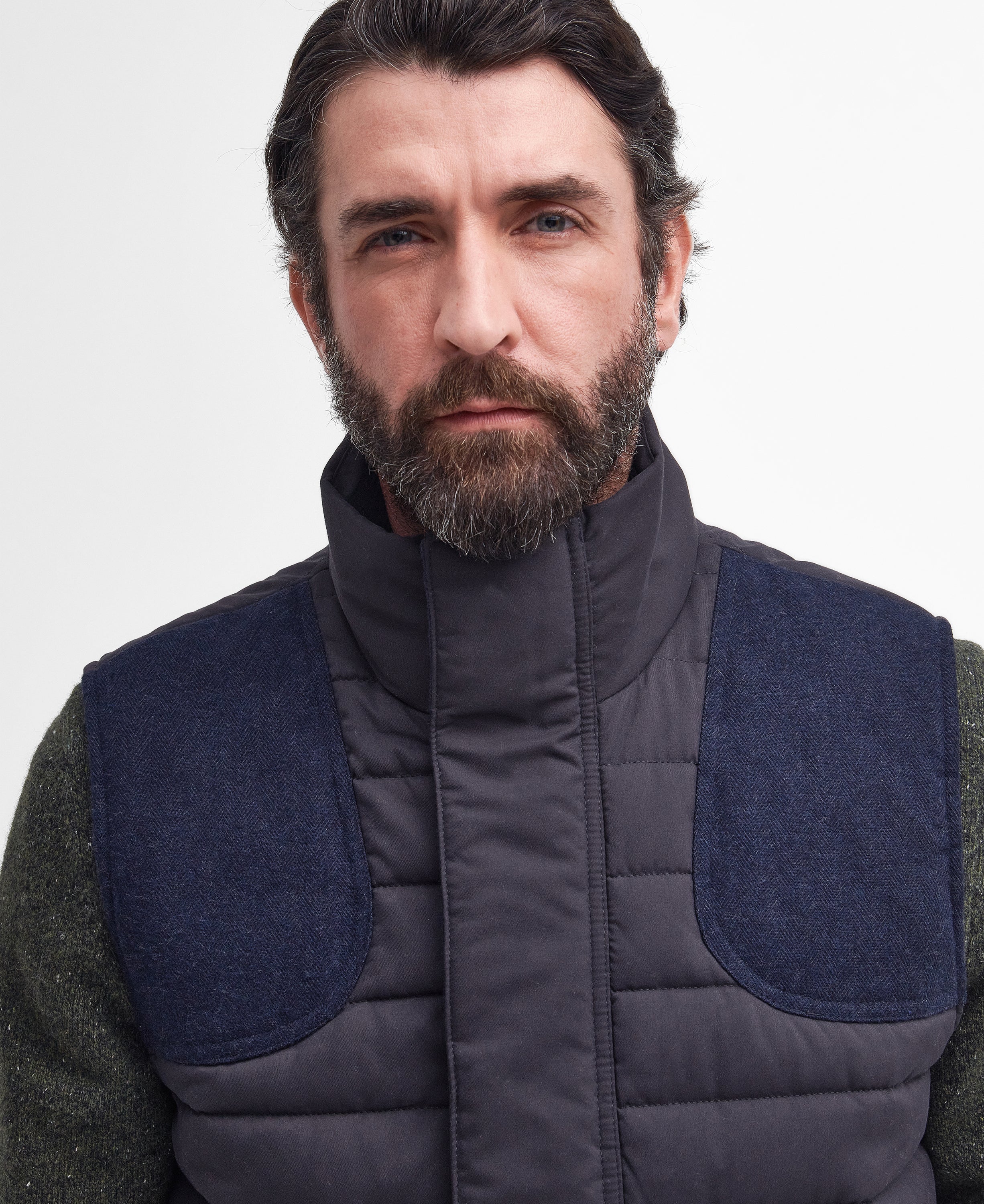 A close-up of a man wearing a dark gray quilted puffer vest with navy blue contrast shoulder panels and a high collar, over a green knit sweater, against a plain light gray background.