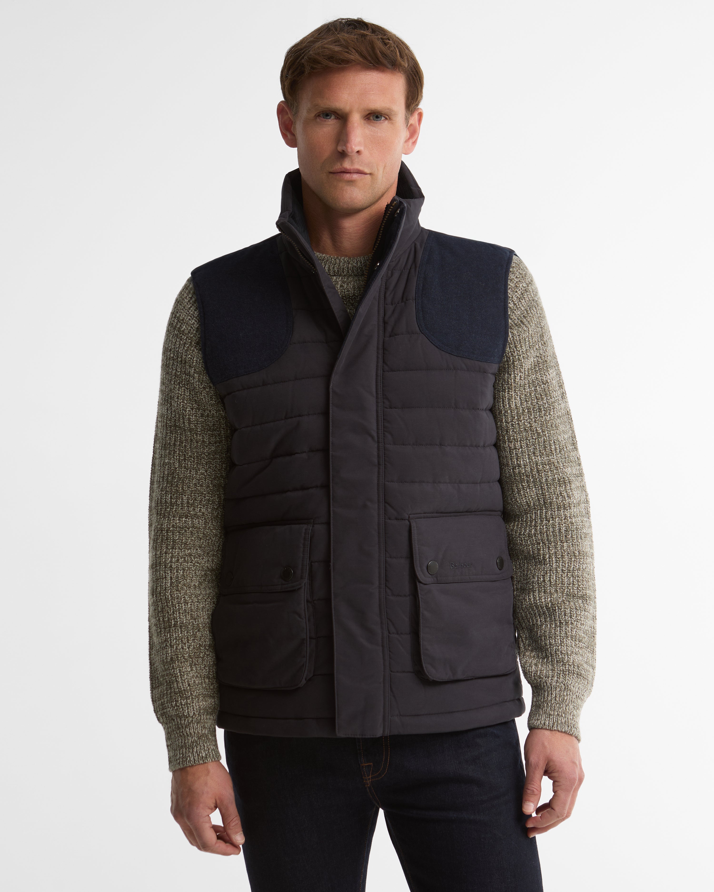 A man wearing a dark navy quilted puffer vest over a textured olive green knit sweater and dark jeans, photographed against a clean white studio background.