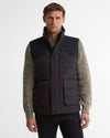 A man wearing a dark navy quilted puffer vest over a textured olive green knit sweater and dark jeans, photographed against a clean white studio background.