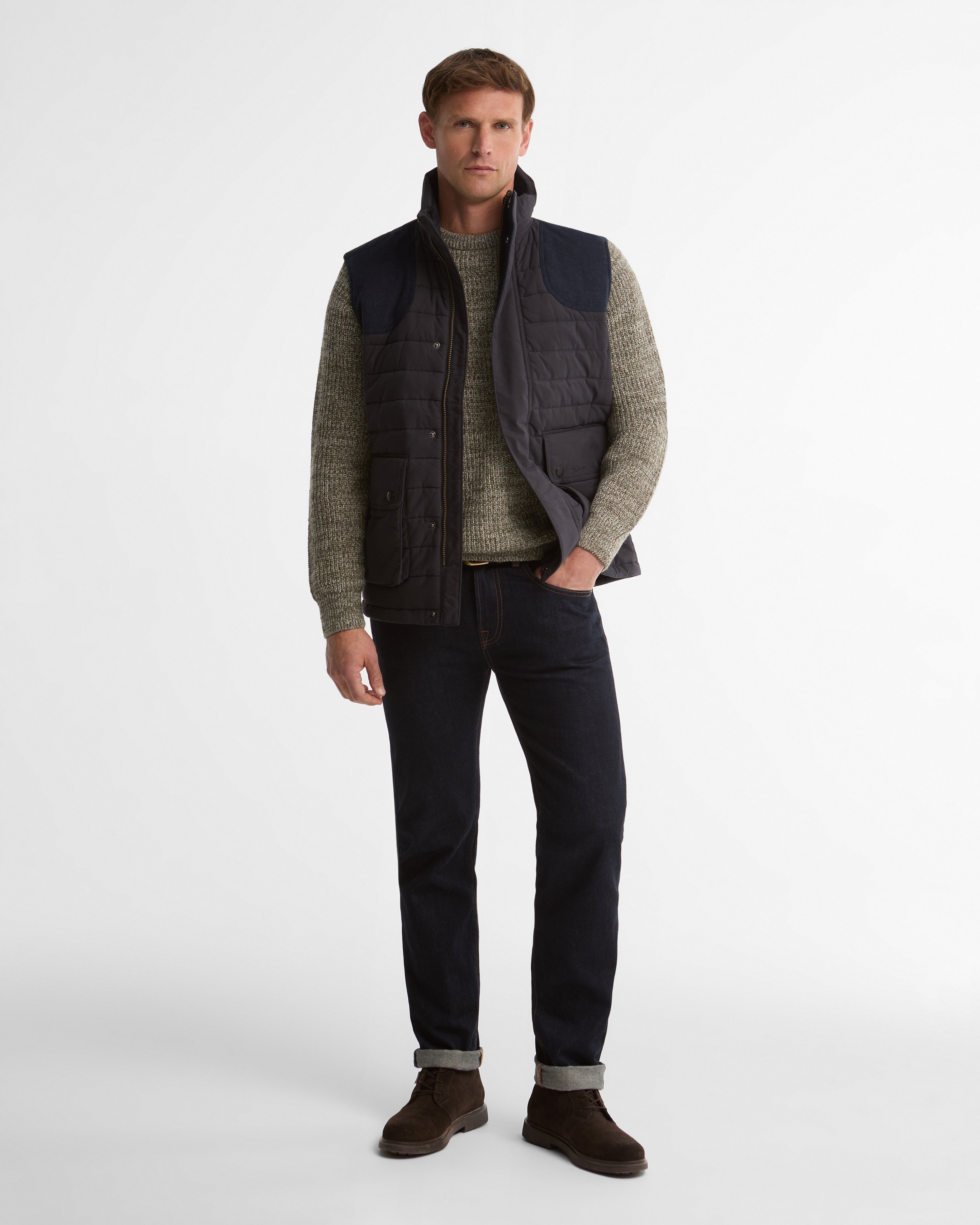 A man modeling a dark quilted sleeveless puffer vest over a textured gray sweater, dark jeans, and brown boots against a plain light gray studio background.