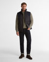 A man modeling a dark quilted sleeveless puffer vest over a textured gray sweater, dark jeans, and brown boots against a plain light gray studio background.